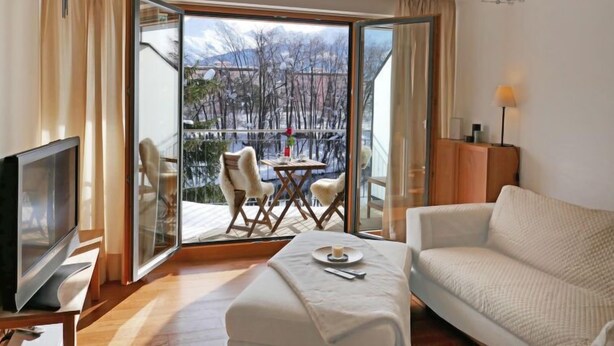 Ein modernes Hotelzimmer mit einer großen Schiebeglasdoor zum Balkon mit Blick auf Bäume und Berge. Es gibt einen Flachbildfernseher, eine Sitzecke und einen kleinen Tisch mit Kerze und Fernbedienung.