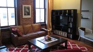 A living room with a leather sofa, a coffee table, a bookshelf, and a large window with curtains. - Modern meets Historic, Natural Lighting + High Ceilings, Near Metro (Belleville)