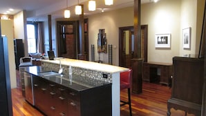 A modern kitchen with a central island, wooden flooring, pendant lighting, and a view of the living area. - Modern meets Historic, Natural Lighting + High Ceilings, Near Metro (Belleville)