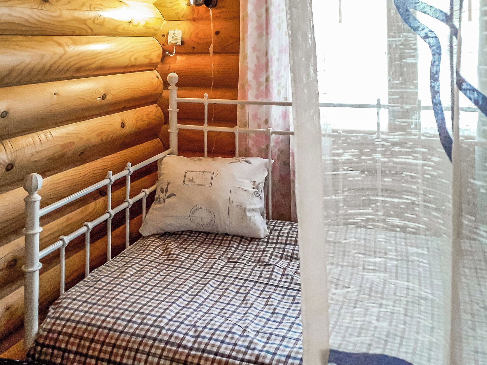 A wooden cabin room with a metal bed frame, a checkered bedspread, and a single pillow.