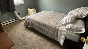 Un dormitorio con cama, calefactor, ventana con cortinas y piso alfombrado.