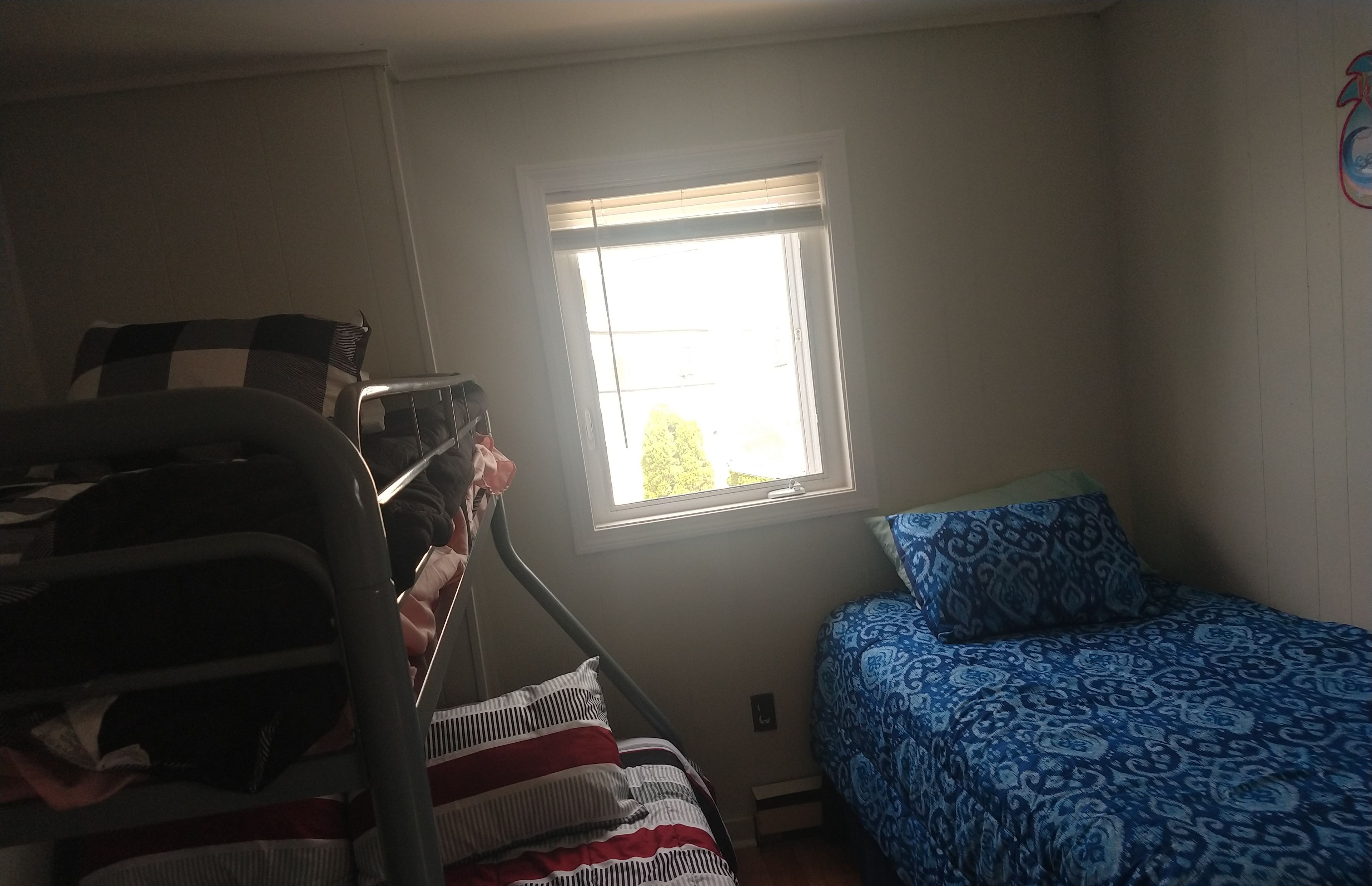 A bedroom with a bunk bed, a single bed with patterned bedding, a window with blinds, and a wall-mounted shelf.