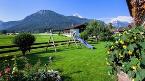 Un terrain de jeux avec un toboggan et des balançoires, entouré d’une pelouse verdoyante et de plantes en fleurs.