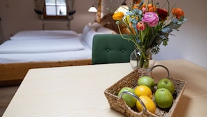 Ein Zimmer mit einem Bett, einem grünen Sessel, einem Tisch mit Obstkorb und einer Vase mit Blumen.