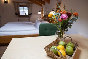 A room with a bed, a green chair, a table with a fruit basket, and a vase with flowers.