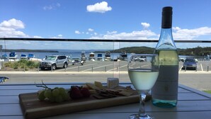 A table with a bottle of white wine, a glass, and a wooden board with grapes and cheese. A marina with boats and cars is visible in the background. - Swansea Lakeside Retreat is a large property on the shores of Lake Macquarie (Swansea)
