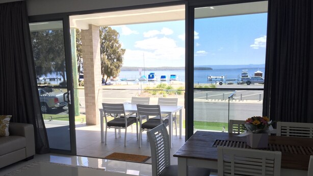 A modern living room with a large sliding glass door leading to a balcony. The room features a white sofa, a dining table with chairs, and a small coffee table. - Swansea Lakeside Retreat is a large property on the shores of Lake Macquarie (Swansea)