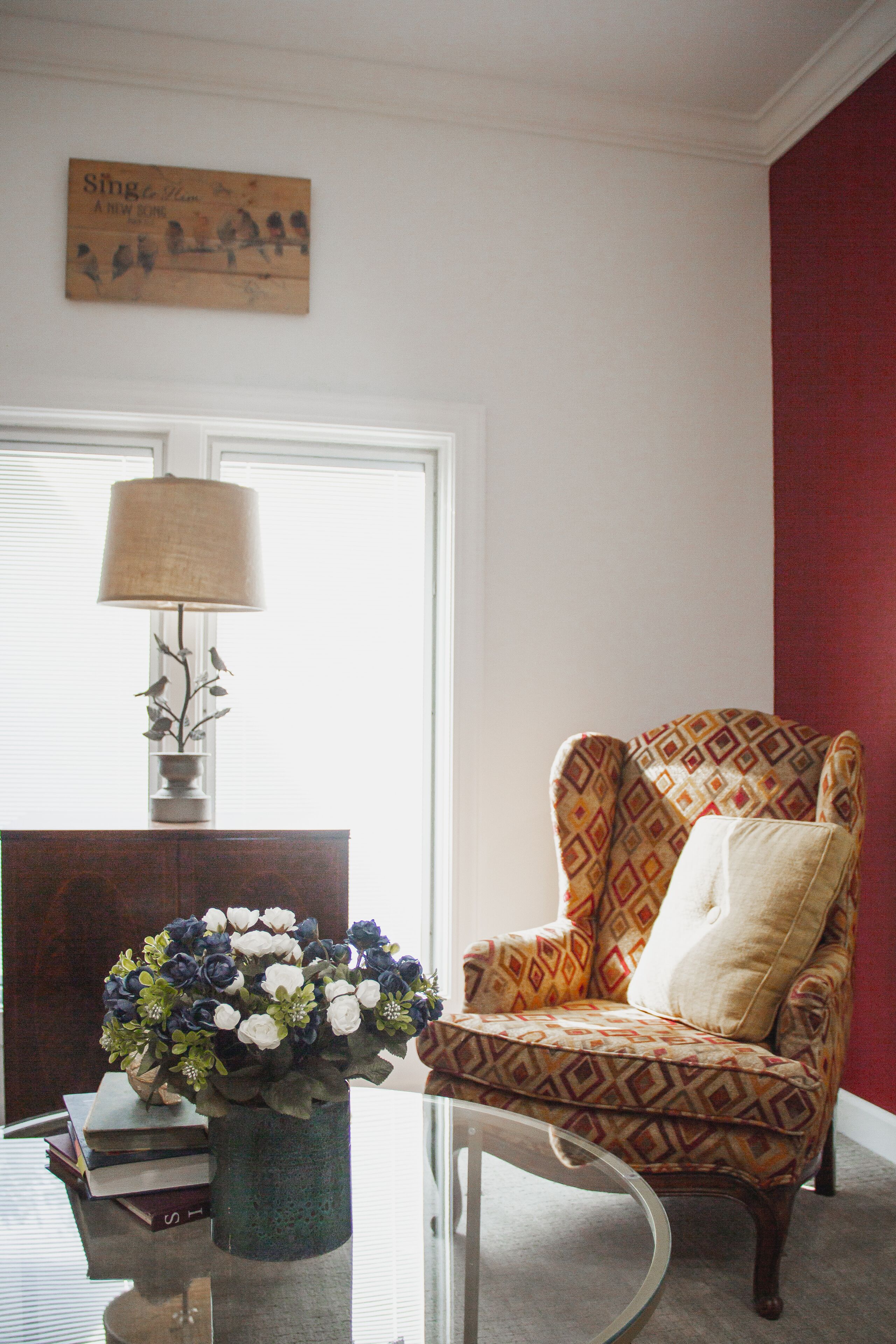 Una sala de estar con un sillón estampado, un florero con flores, una lámpara y un cuadro en la pared.