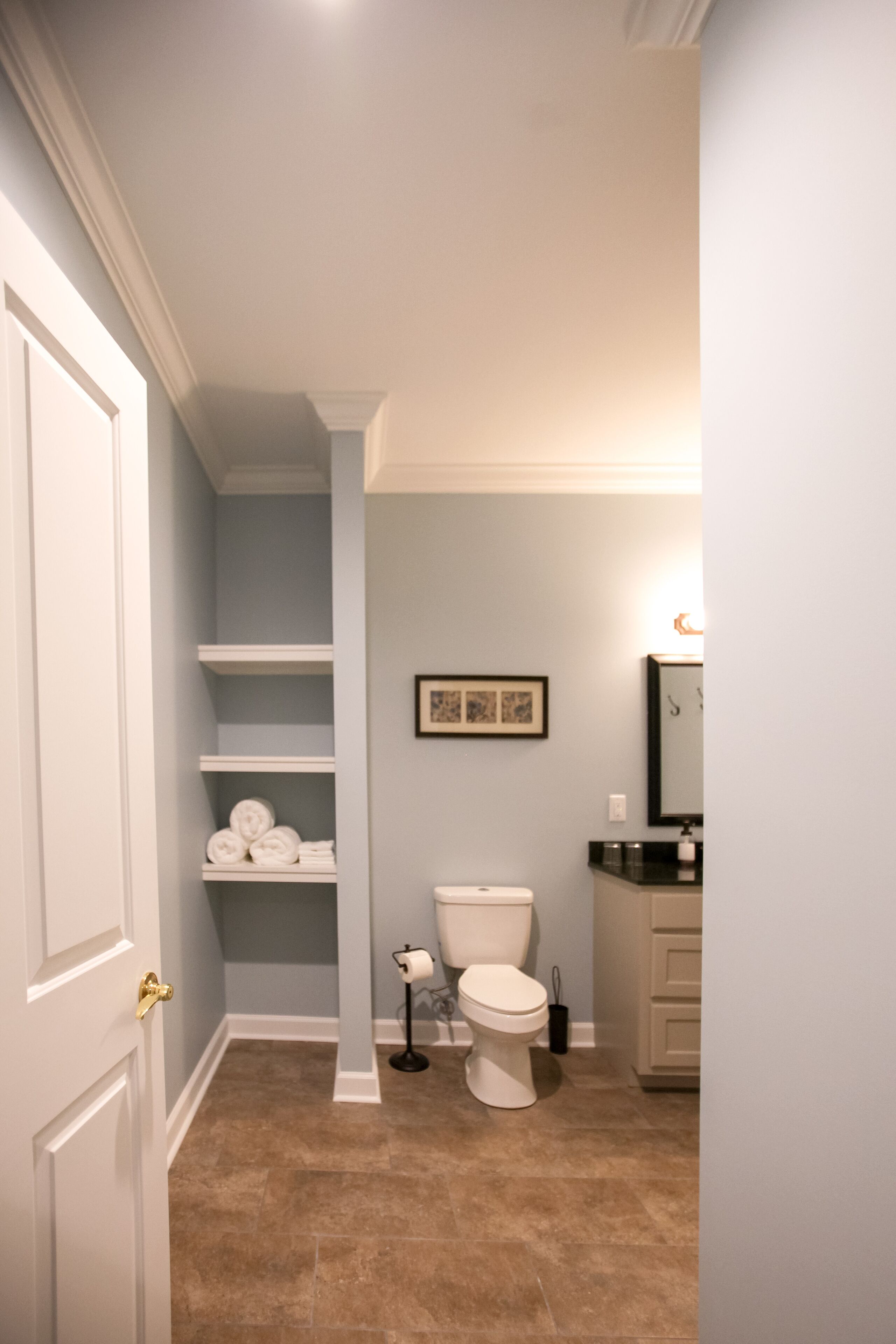 A bathroom with a toilet, shelves with towels, a framed picture on the wall, and a sink with a mirror.