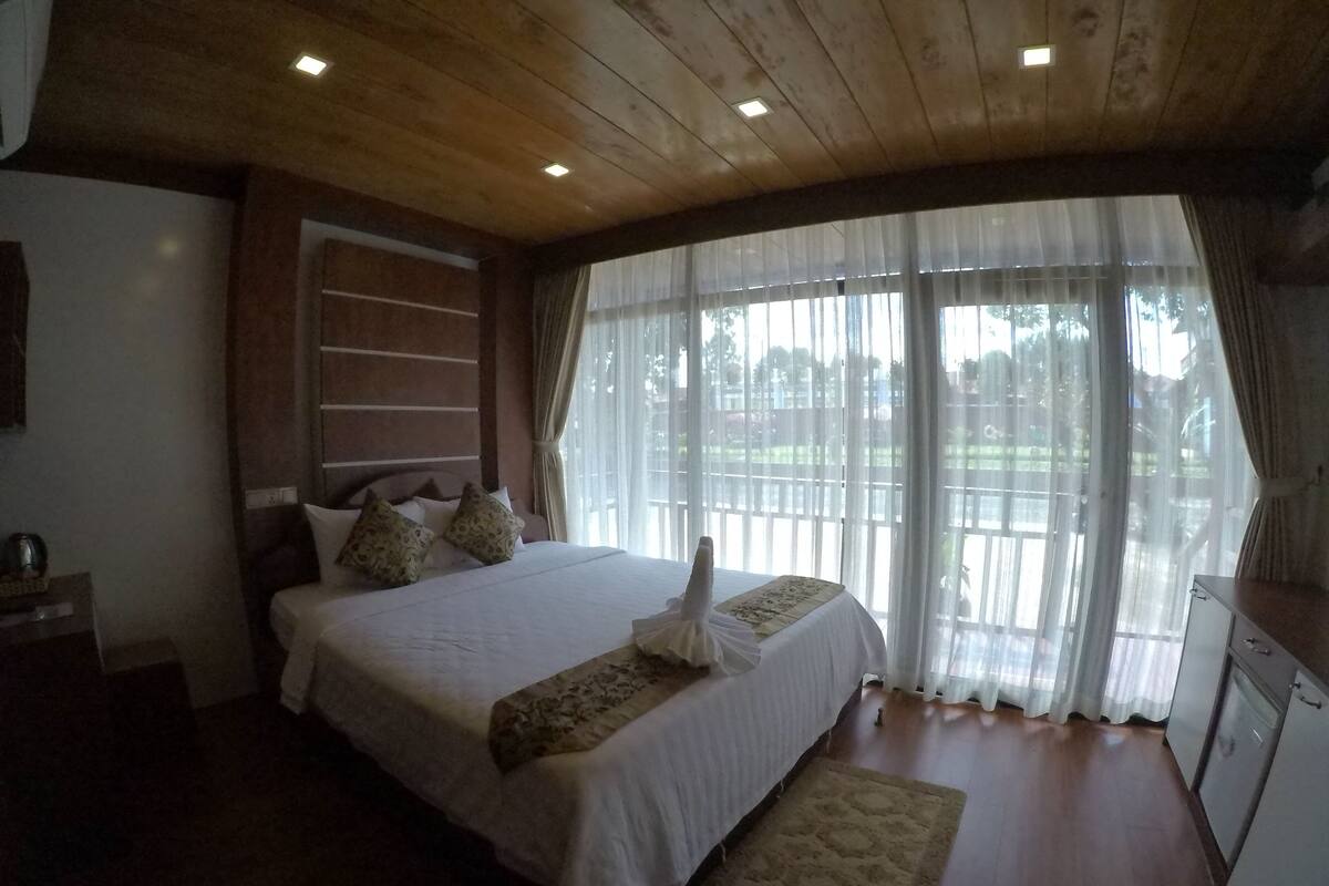 A bedroom with a wooden ceiling, a large bed with white bedding, a bedside table, and a view through sheer curtains.