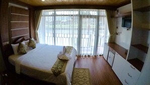 Quarto com teto de madeira, cama com roupa de cama branca, TV na parede e uma janela grande com cortinas.