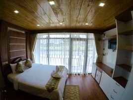 Une chambre avec un plafond en bois, un lit recouvert de draps blancs, un téléviseur fixé au mur et une grande fenêtre avec des rideaux.