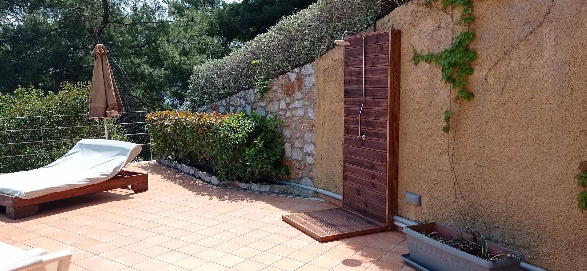 Un espacio al aire libre iluminado por el sol, con un banco de madera, una silla lounge cubierta y una puerta de madera, junto a una pared de piedra con plantas.