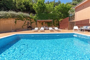 A swimming pool with lounge chairs, a covered seating area, and a building with a wooden fence and greenery.