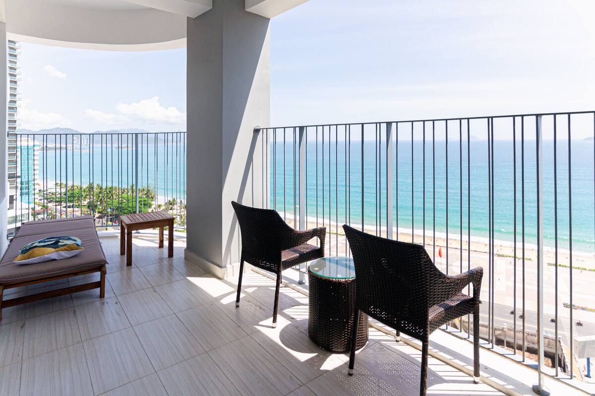 Een balkon met uitzicht op het strand, twee rieten stoelen, een klein tafeltje en een bed met een kussen.