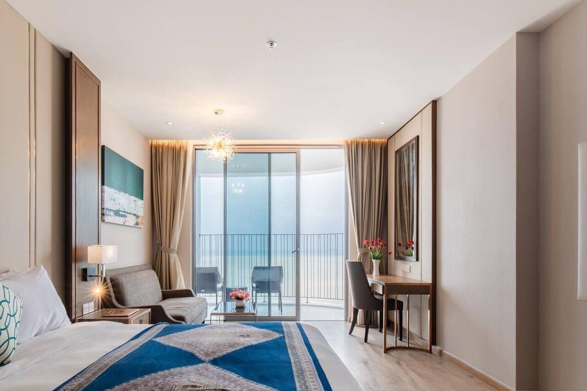Hotelkamer met een groot bed, een bureau, een stoel, een klein tafeltje met bloemen en uitzicht op zee door een schuifdeur.