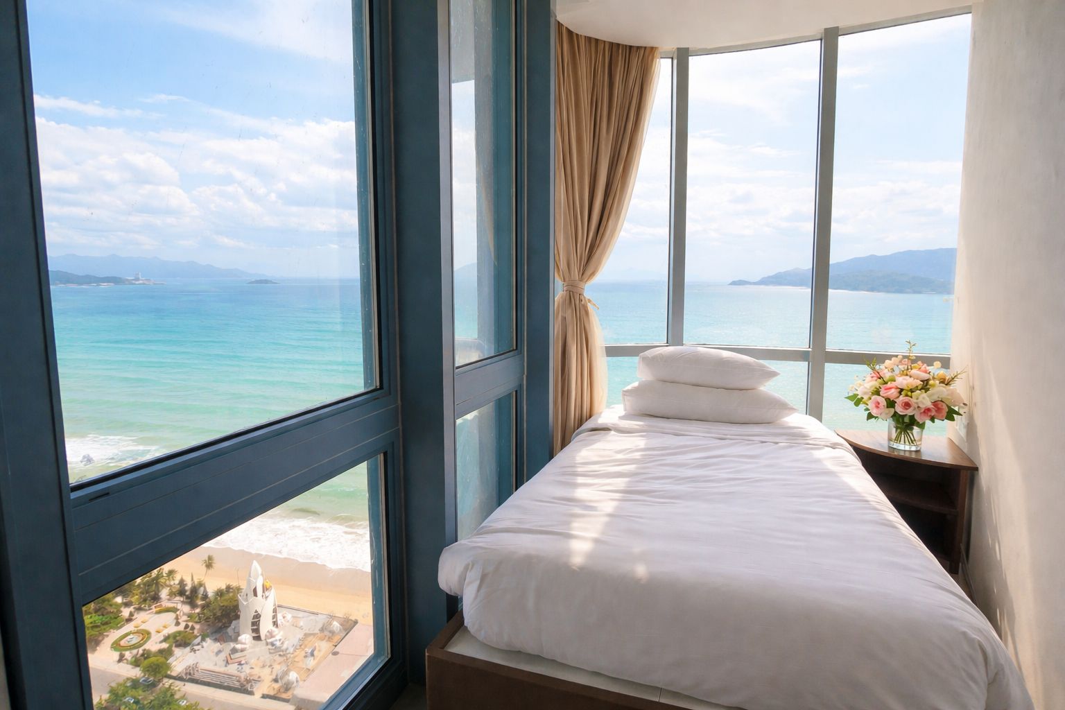 A hotel room with a bed, a window with a view of the sea, and a vase with flowers on a nightstand.
