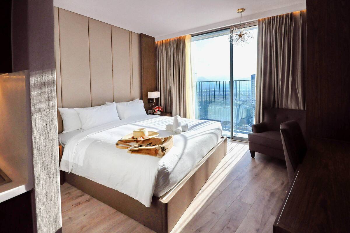 Een hotelkamer met een groot bed, een stoel, een nachtkastje met lamp en uitzicht op de stad.
