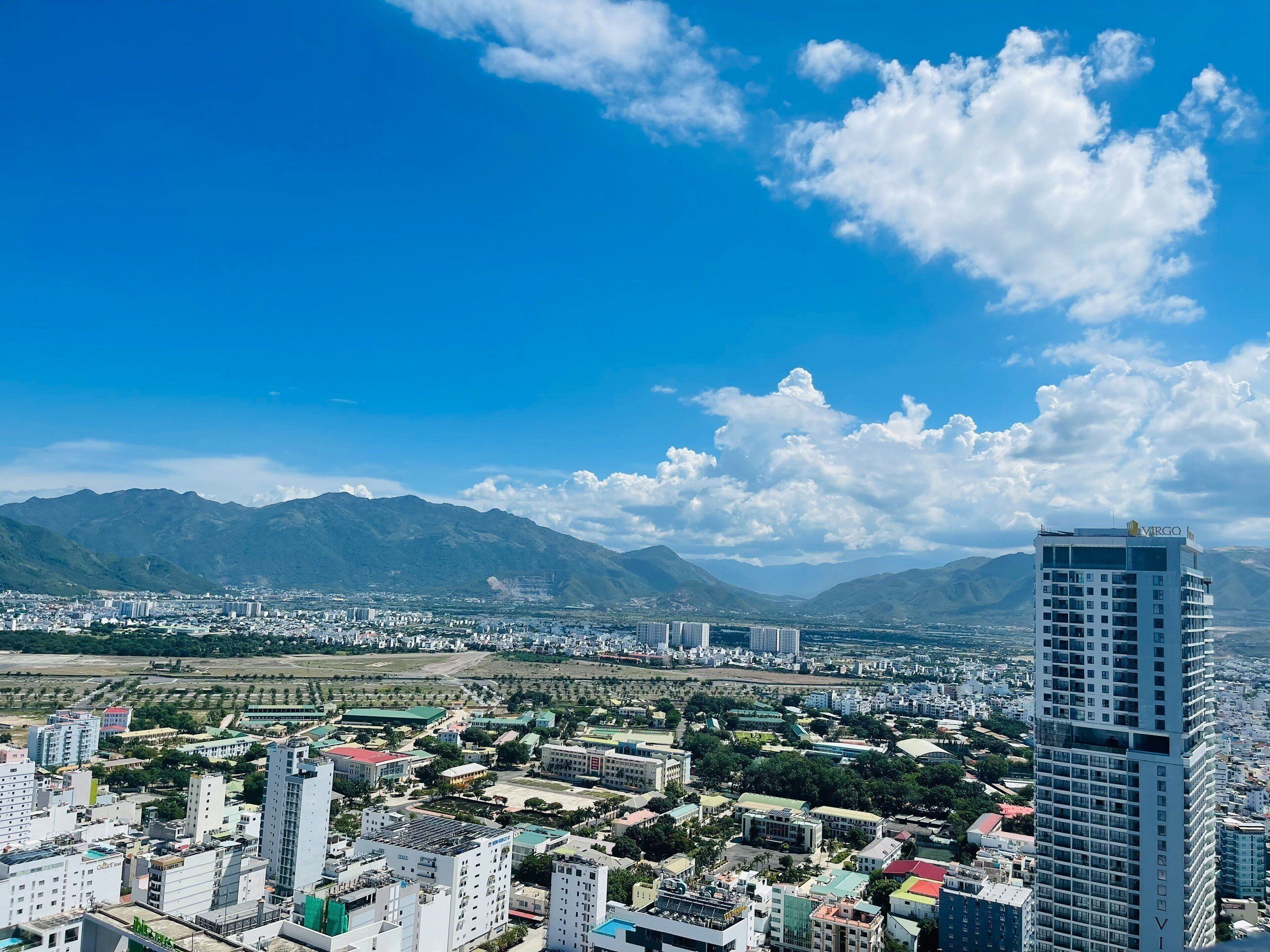 A cityscape with a prominent modern high-rise hotel, surrounded by residential buildings and a mountainous backdrop.