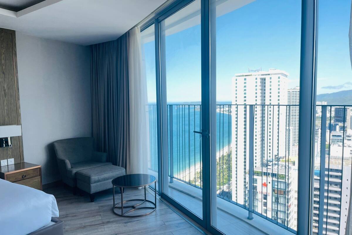 Een moderne hotelkamer met een grote schuifglazen deur naar een balkon, een grijze bank, een ronde tafel en een bed met witte lakens.