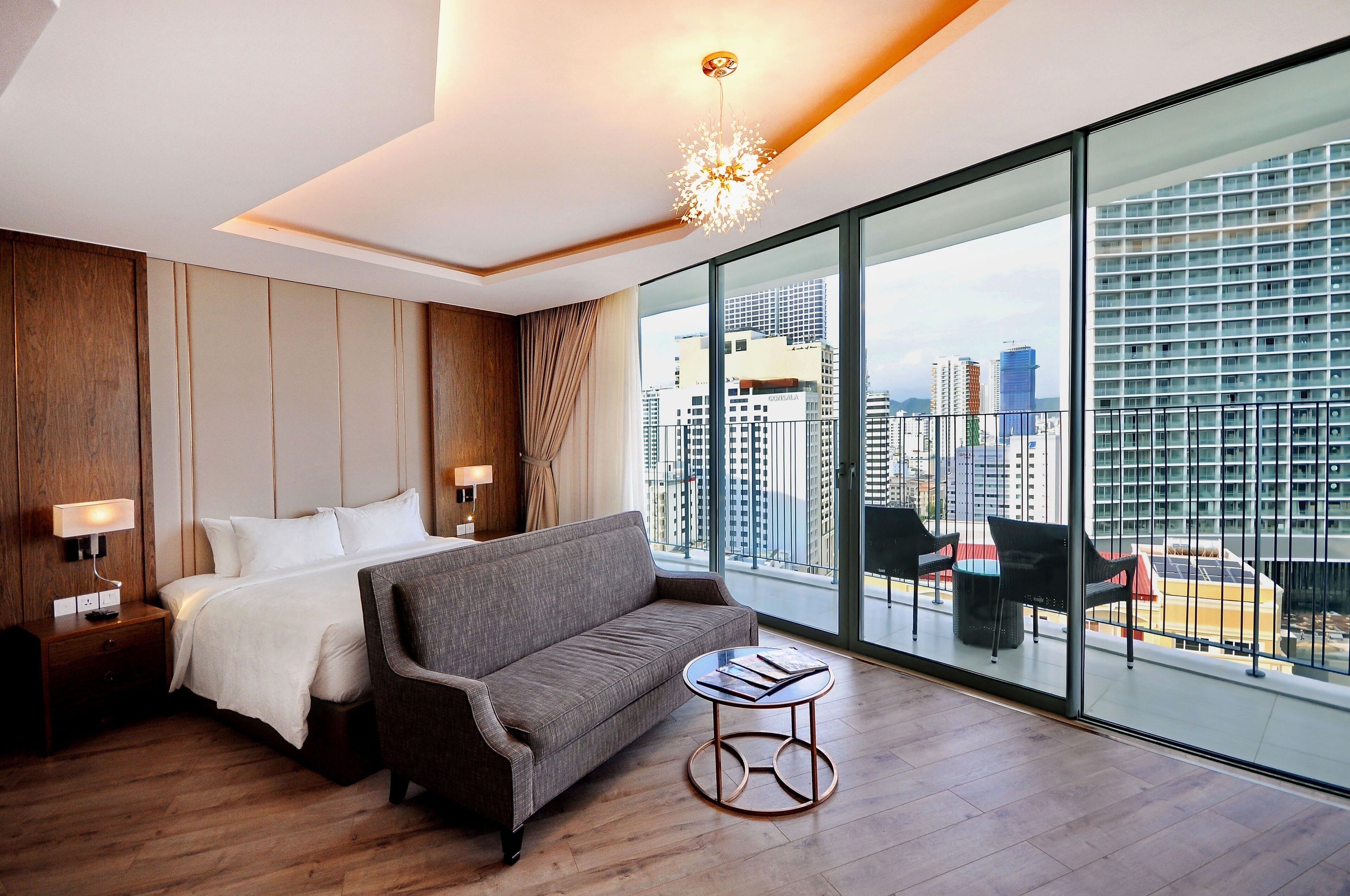 A hotel room with a bed, a sofa, a small table, and a large window offering a city view.