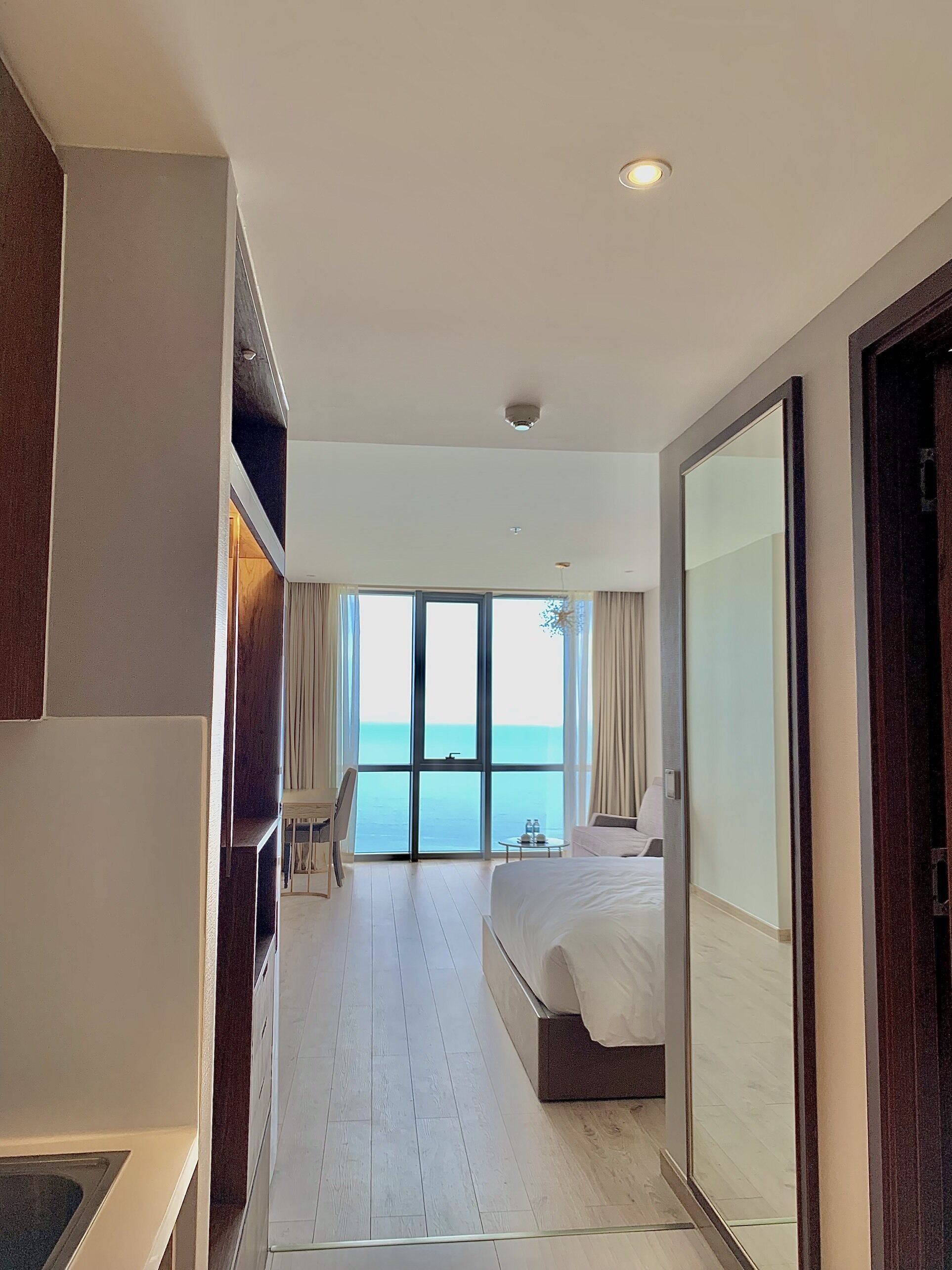 A modern hotel room with a large bed, a seating area with a table, and a view of the sea through a large window.