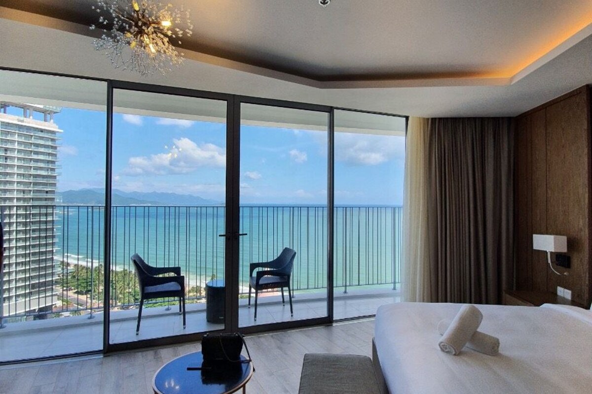 A modern hotel room with a large balcony, a bed, two chairs, a small table, and a view of the ocean and a high-rise building.