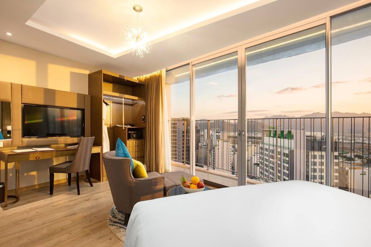 Een hotelkamer met een groot bed, een bureau met stoel, een televisie en een balkon met uitzicht over de stad.