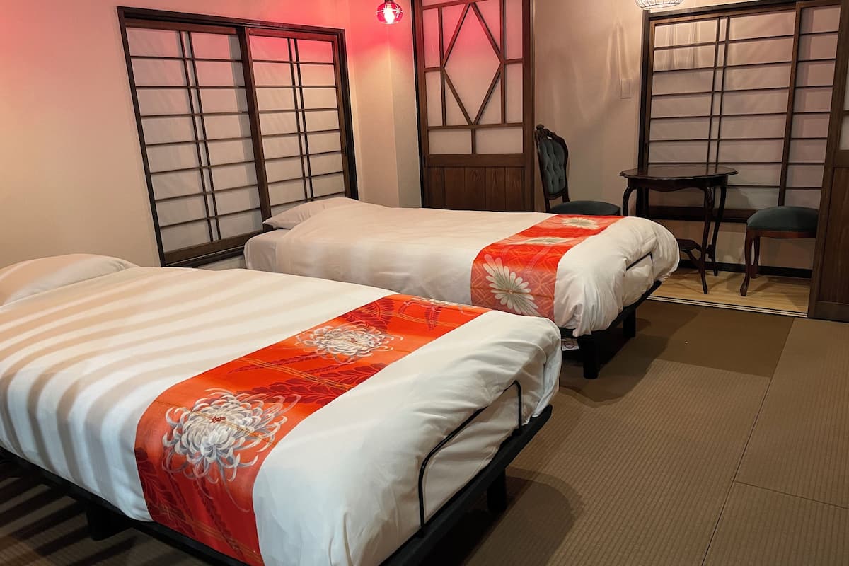 Deux lits avec une literie blanche et orange, une chaise et une petite table dans une pièce dotée de portes coulissantes de style japonais traditionnel et d’un plancher en bois.