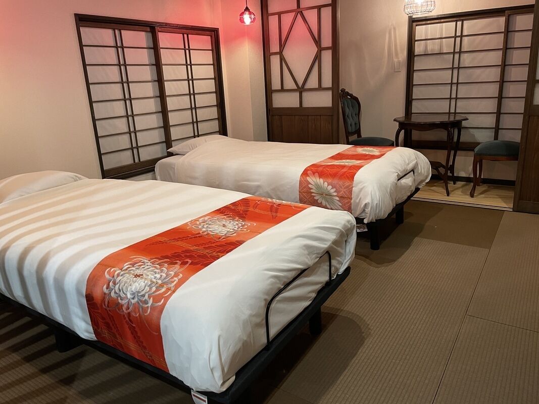 A hotel room with two beds, a chair, a small table, and traditional sliding doors.
