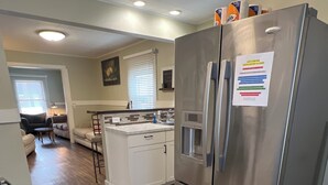 A modern kitchen with a stainless steel refrigerator, a built-in oven, and a sink. There is a small island with a bar stool and a window with blinds. The floor is wooden, and there is a rug in front of the door.