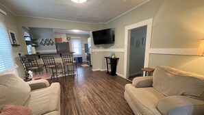 A living room with two sofas, a coffee table, and a TV mounted on the wall. There is a kitchen area in the background with a dining table and chairs.