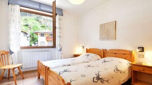 A bedroom with a wooden bed, a chair, a nightstand, a window with curtains, and a view of buildings outside.