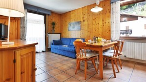 A cozy room with a wooden wall, a blue sofa, a dining table with chairs, a lamp, and a window with a view.