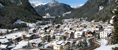 A snow-covered mountain village with traditional chalet-style buildings, a winding road, and a backdrop of snow-capped peaks.