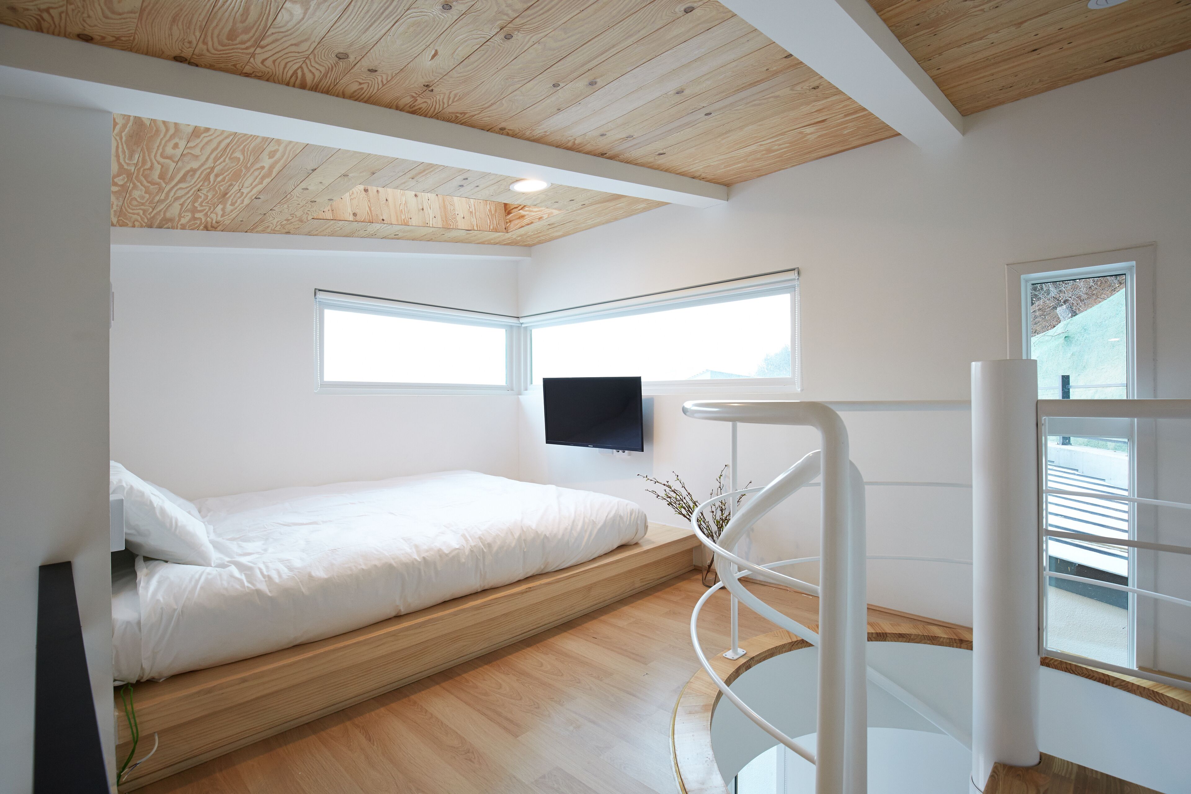 A modern bedroom with a wooden ceiling, a bed, a TV mounted on the wall, and a spiral staircase.