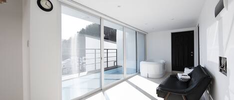 Una habitación moderna y luminosa con un gran ventanal corredero que da a un balcón, una butaca de cuero negro y un banco blanco.