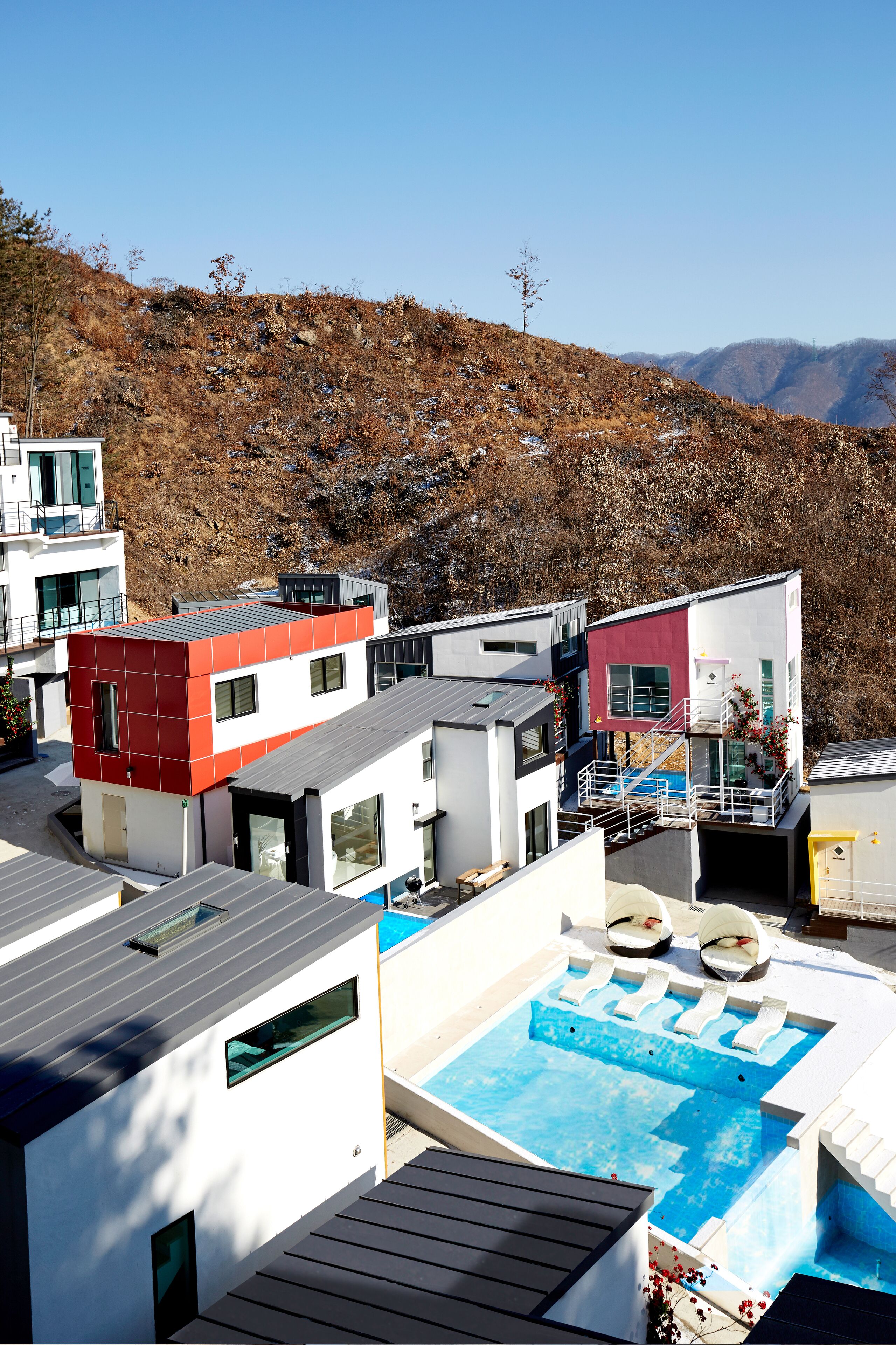 A modern residential complex with a swimming pool, multiple buildings, and a hillside in the background.