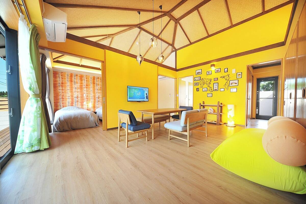 A bright yellow room with wooden floors, a bed, a table with chairs, and a TV on the wall.
