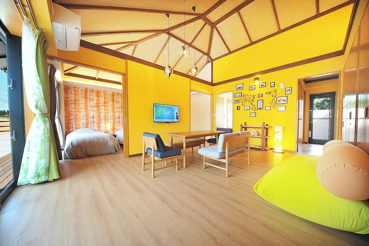 Une pièce lumineuse aux murs jaune vif, dotée d’un plancher en bois, d’un lit, d’une table avec des chaises et d’un téléviseur fixé au mur.
