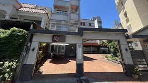 Entrada de um edifício com placa, área de estar coberta ao ar livre e escadaria para um segundo andar.