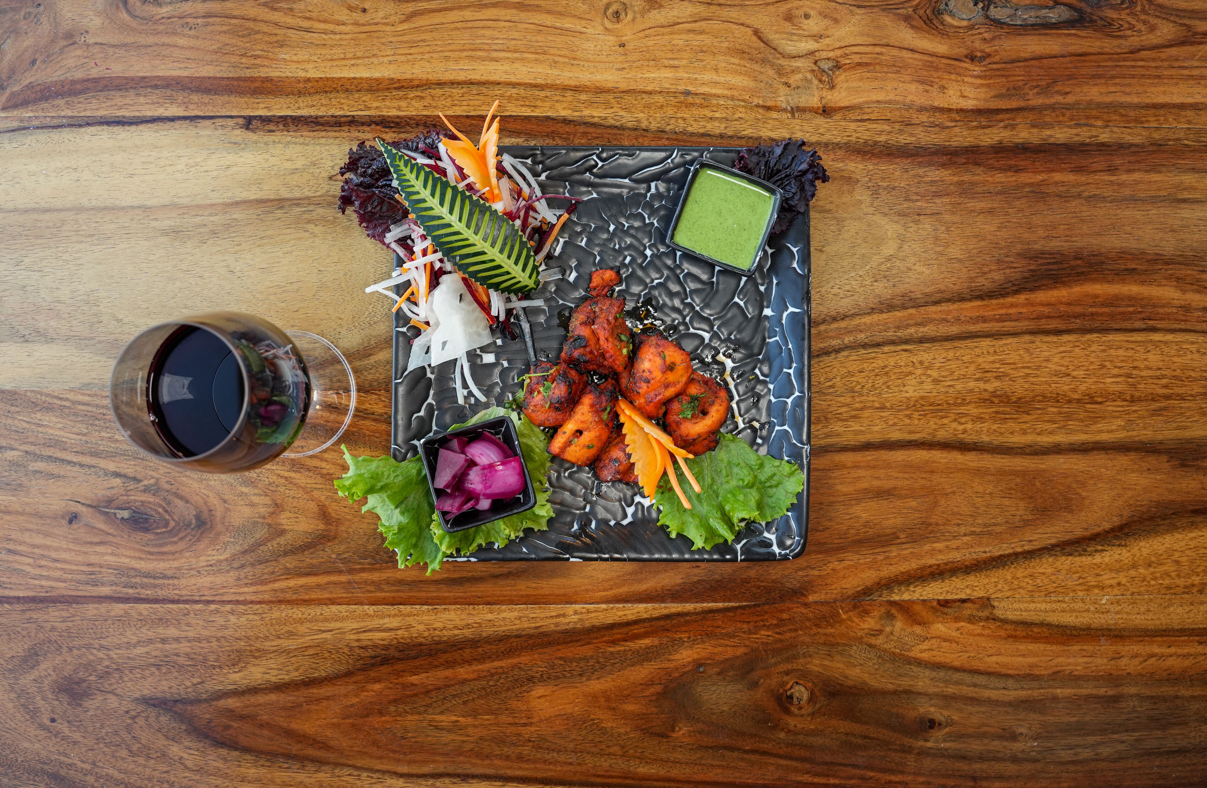 A wooden table with a black plate containing food, a glass of dark liquid, and a small bowl of green sauce.