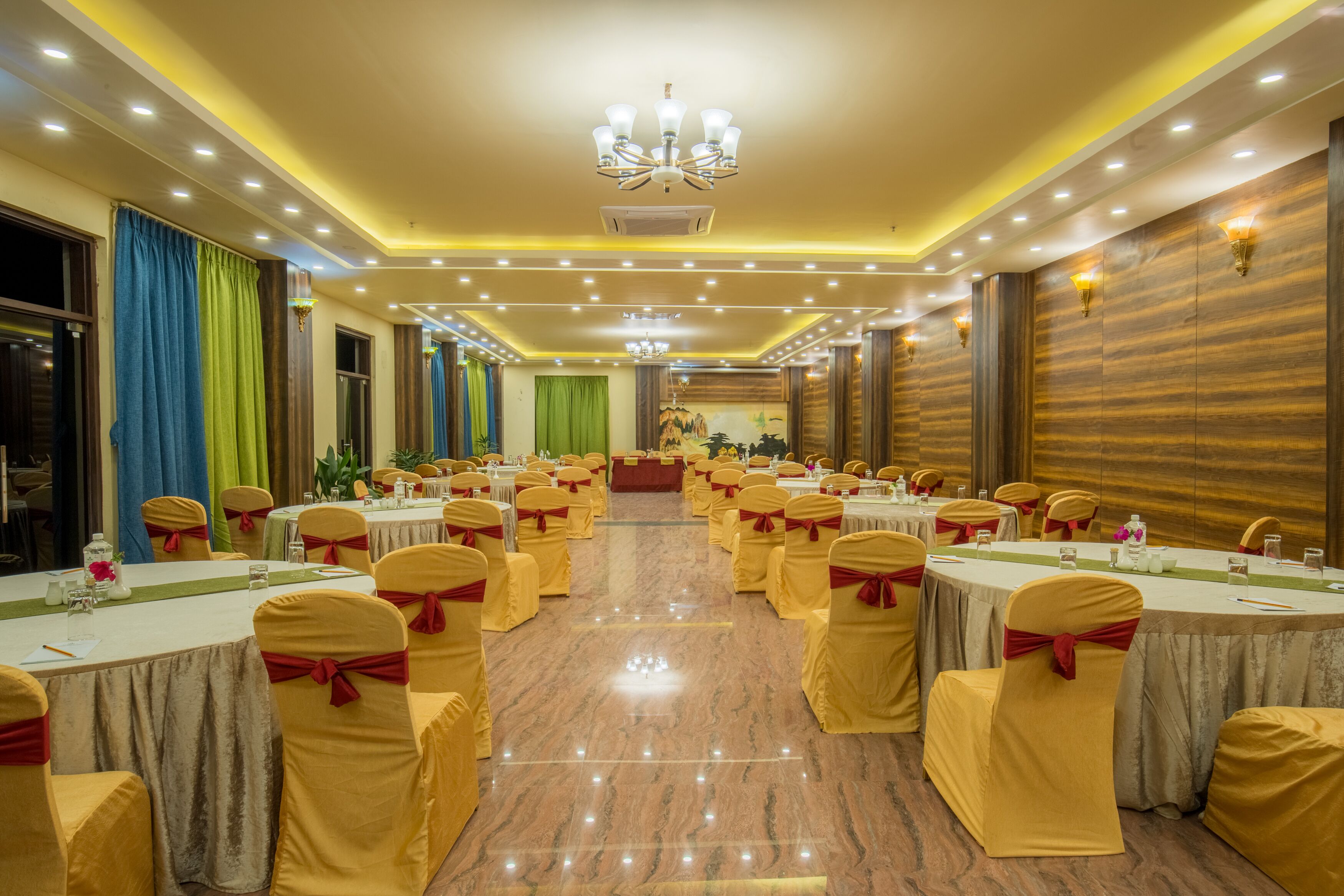 Un salón de banquetes con mesas redondas, sillas amarillas adornadas con lazos rojos y una araña de cristal.