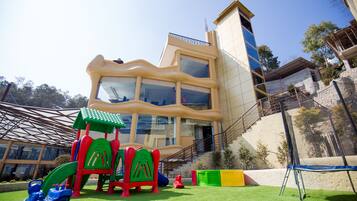 Un parque infantil con toboganes y estructuras para escalar, un edificio con múltiples ventanas y balcones, y un cielo despejado.