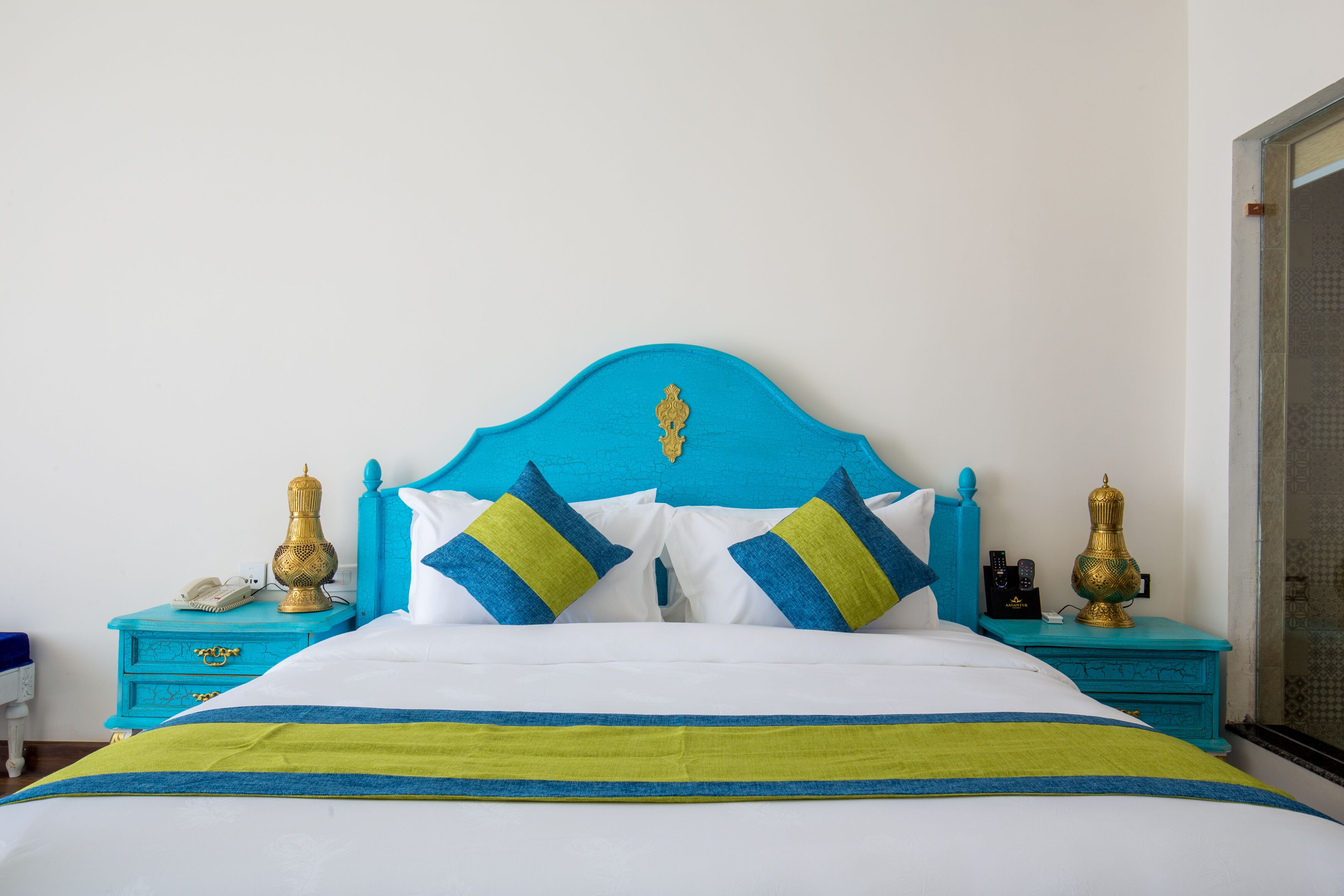 Ein Bett mit blauer Kopfteil, weißen und blauen Kissen und einer grün-blauen Bettdecke. Zwei Nachttische mit Dekoration und ein Telefon auf einem davon.