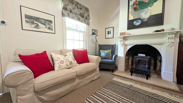 A living room with a beige sofa, red and patterned cushions, a patterned rug, a fireplace, and framed artwork on the walls. - Redruth - 2 bedroom cottage situated in wandiligong (WANDILIGONG)