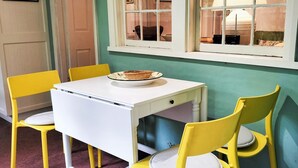 A small dining area with a white table and yellow chairs, a window with a view, and a door in the background. - Redruth - 2 bedroom cottage situated in wandiligong (WANDILIGONG)