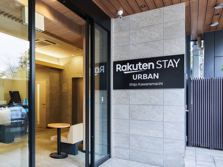 A modern urban hotel entrance with a reception desk, a seating area, and a sign indicating the hotel name 'Rakuten Stay Urban'.