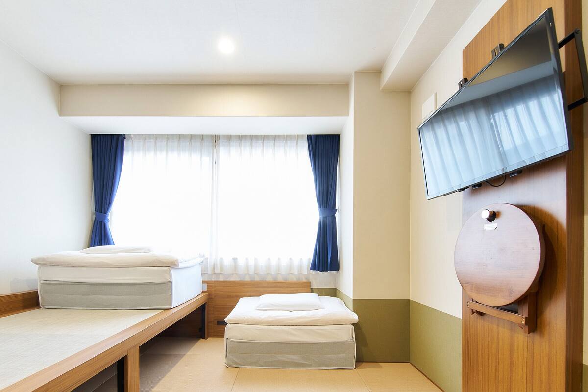 Una habitación pequeña con cama, una mesita, televisor montado en la pared y ventana con cortinas.