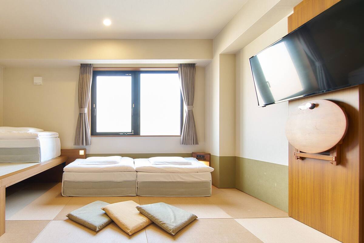 Una habitación con cama, un banco, televisor y ventana con cortinas.